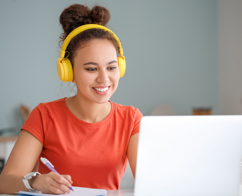 A young woman in an orange shirt with yellow headphones using her white laptop for digital learning at home